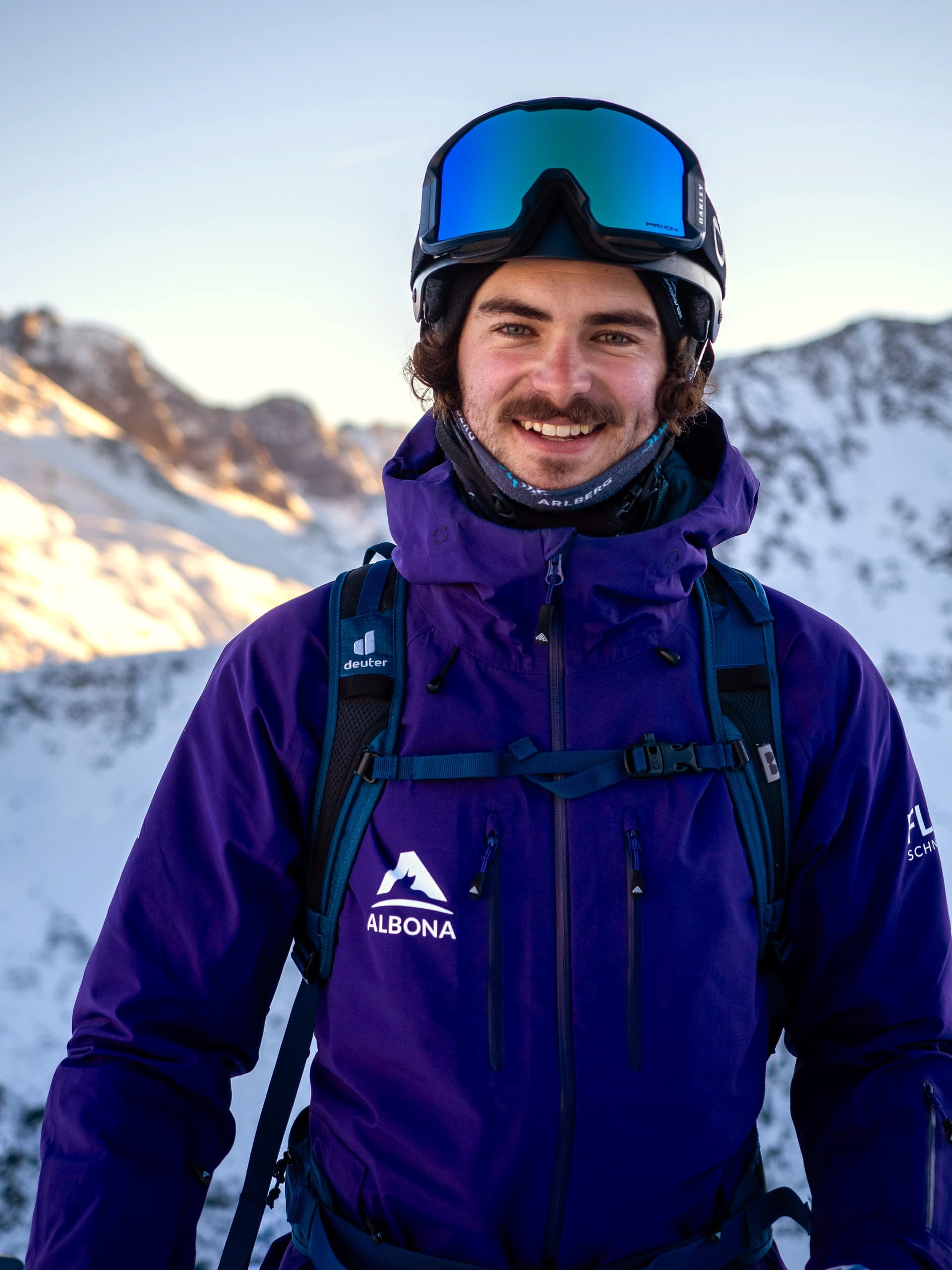 Ein flow Guide in lila Albona-Jacke mit blau verspiegelter Brille lacht in die Kamera, dahinter die sonnigen Gipfel des Arlbergs.