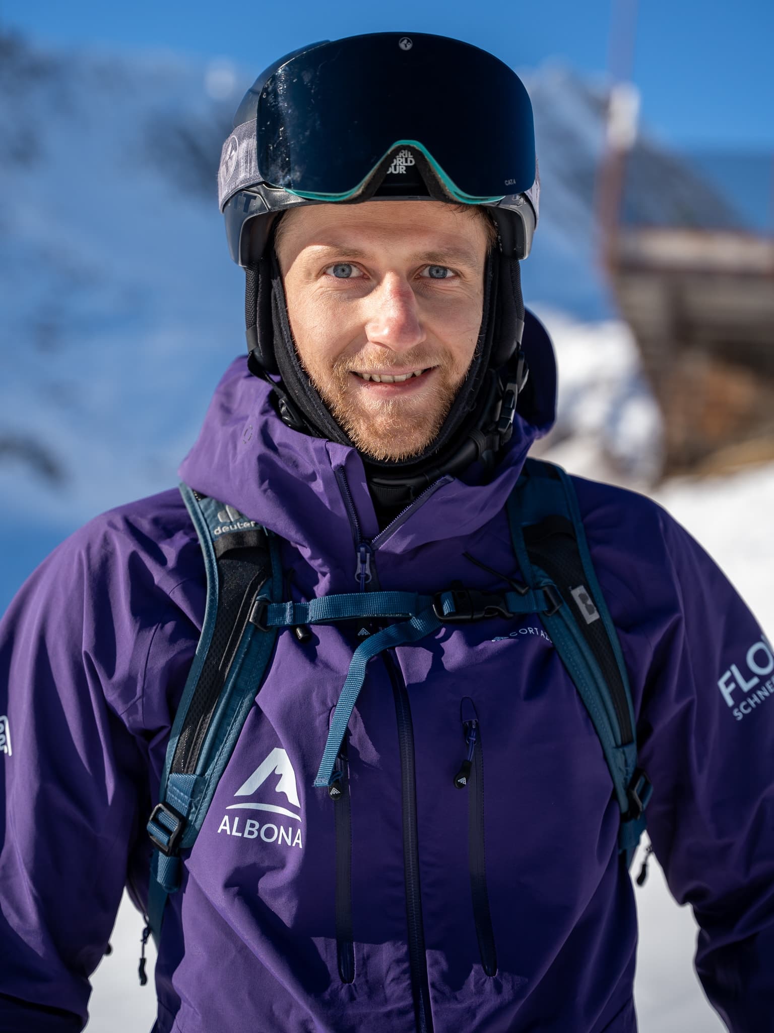 Ein flow Guide in lila Albona-Jacke lächelt vor einer verschneiten Arlberg-Flanke in die Kamera.