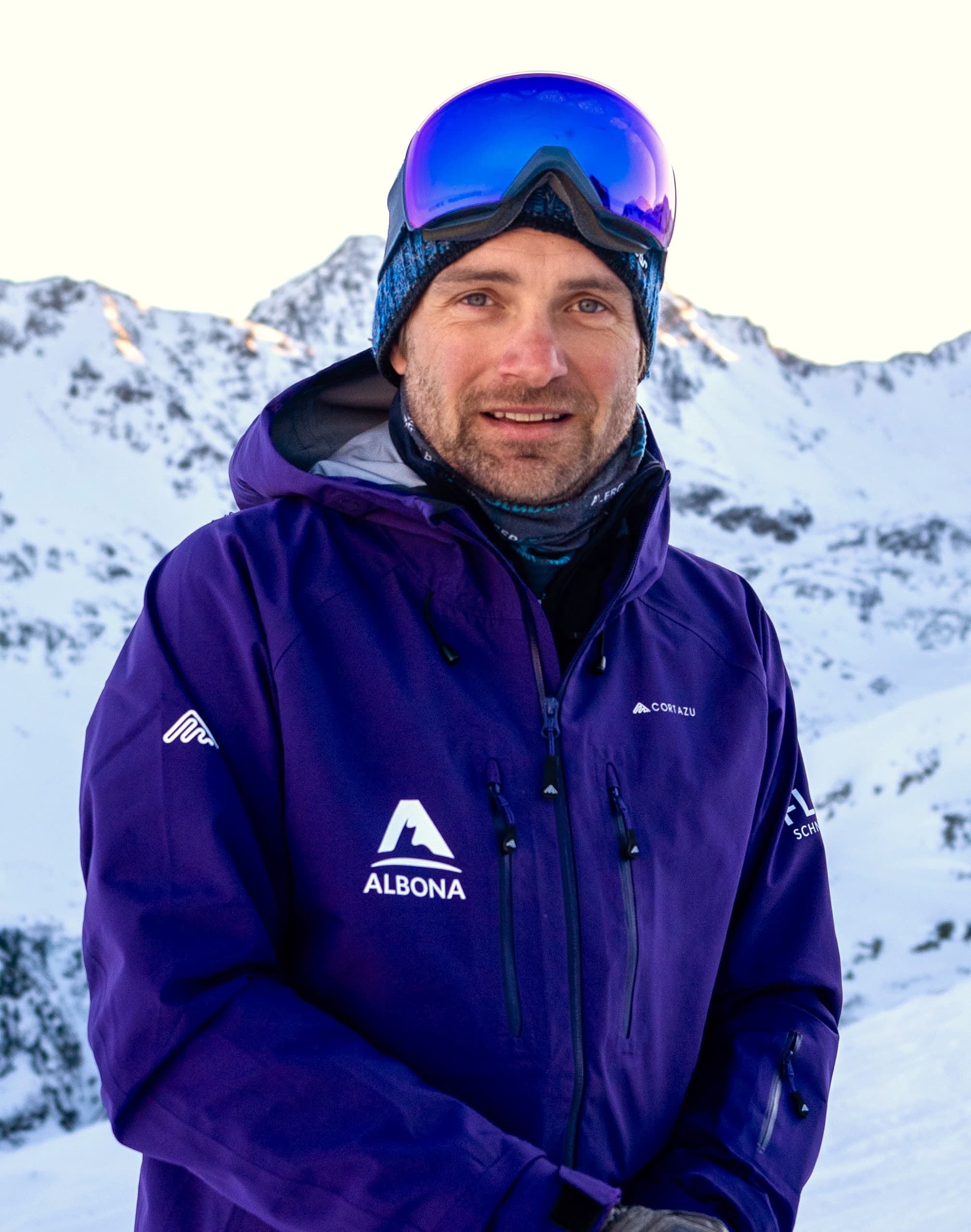 Ein flow Guide in lila Albona-Jacke mit Mütze und blau verspiegelter Brille lächelt vor den verschneiten Bergen des Arlbergs.