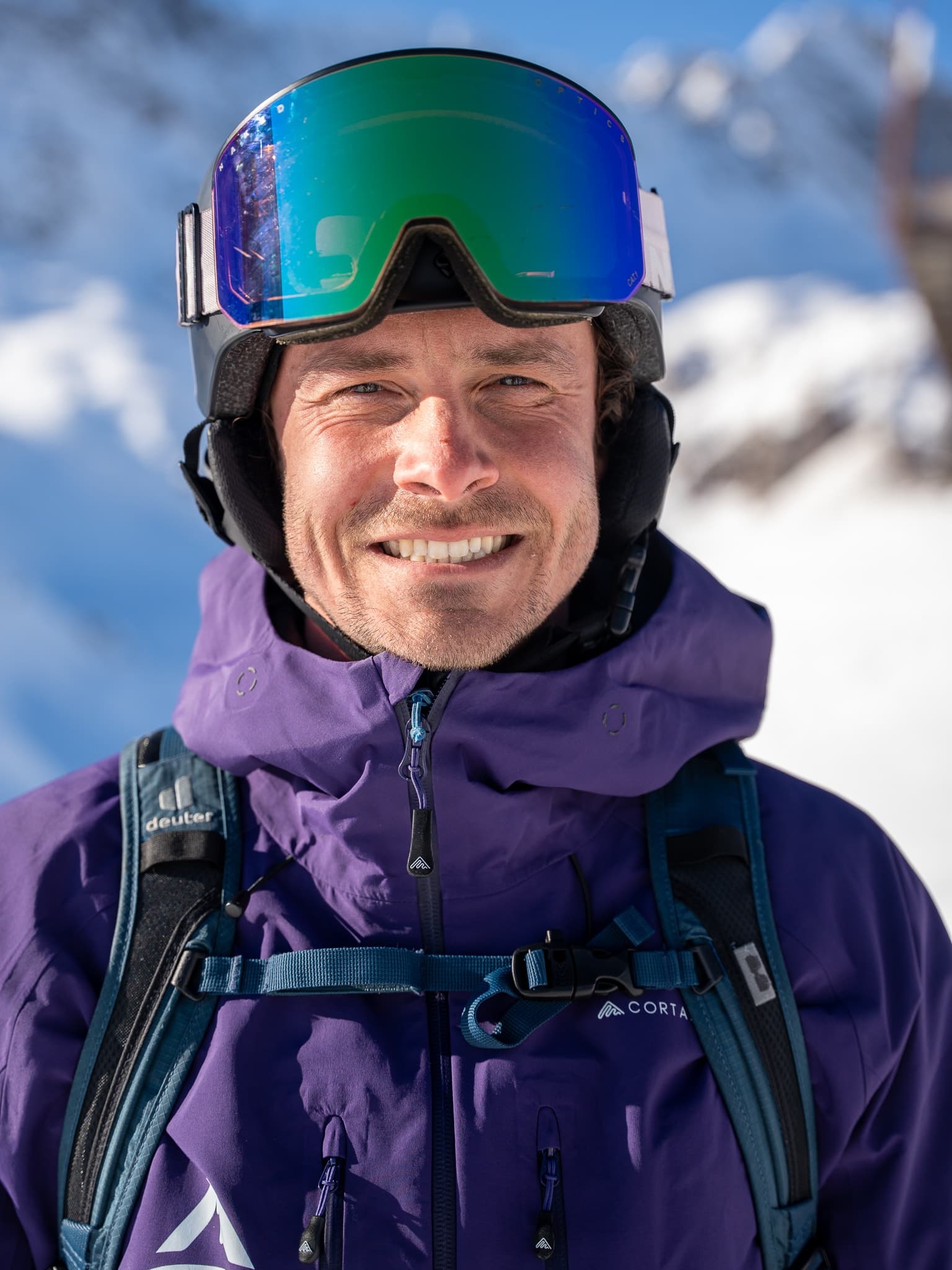Ein flow Guide in lila Albona-Jacke mit grün verspiegelter Brille am Helm, dahinter eine verschneite Arlberg-Flanke.