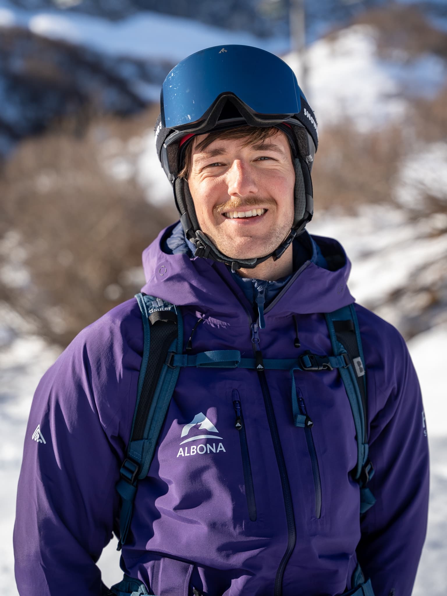 Ein flow Guide in lila Albona-Jacke, die Brille auf den blauen Helm geschoben, lächelt über Stuben am Arlberg in die Kamera.